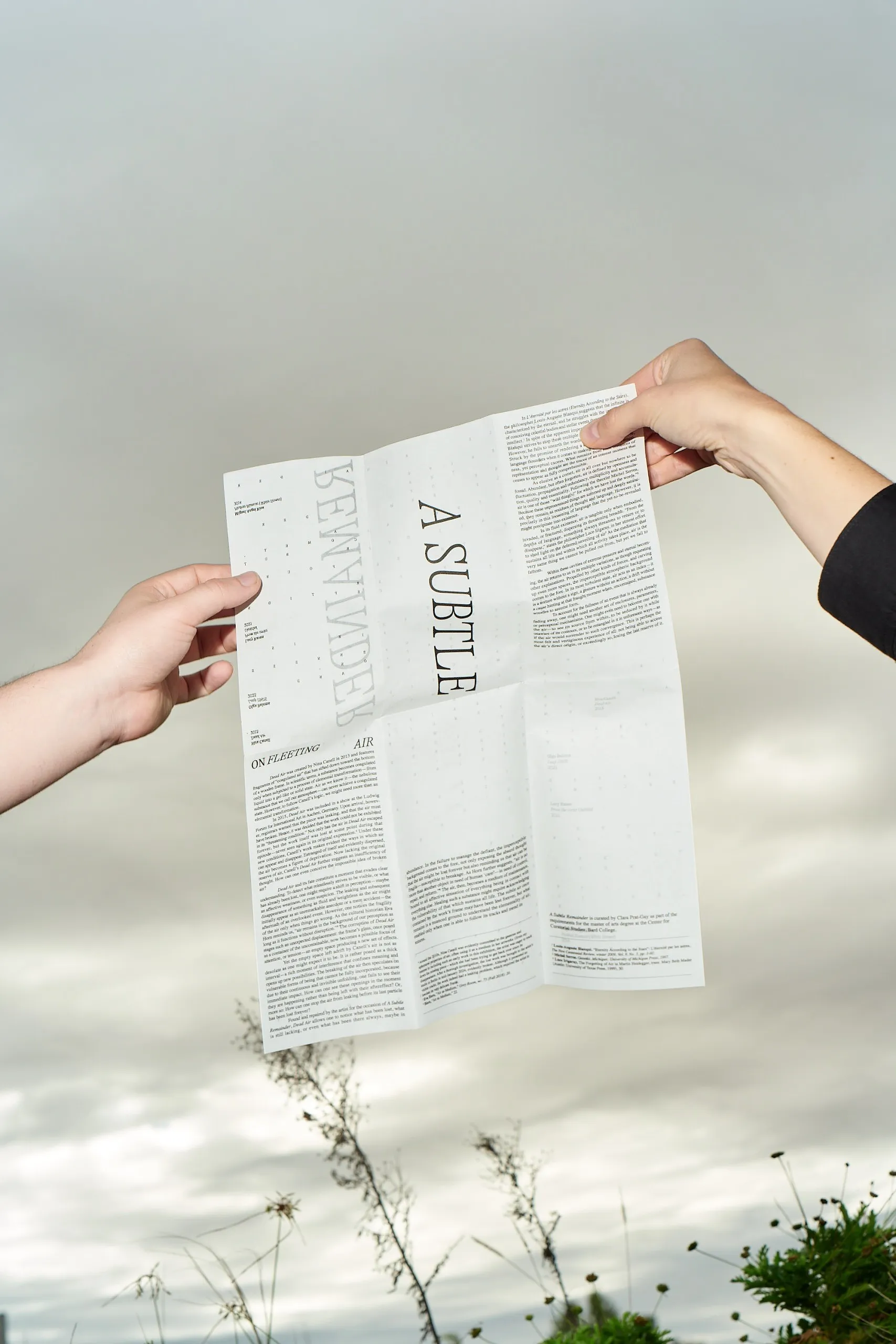 Two hands holding the A Subtle Remainder sheet open outdoors under grey sky