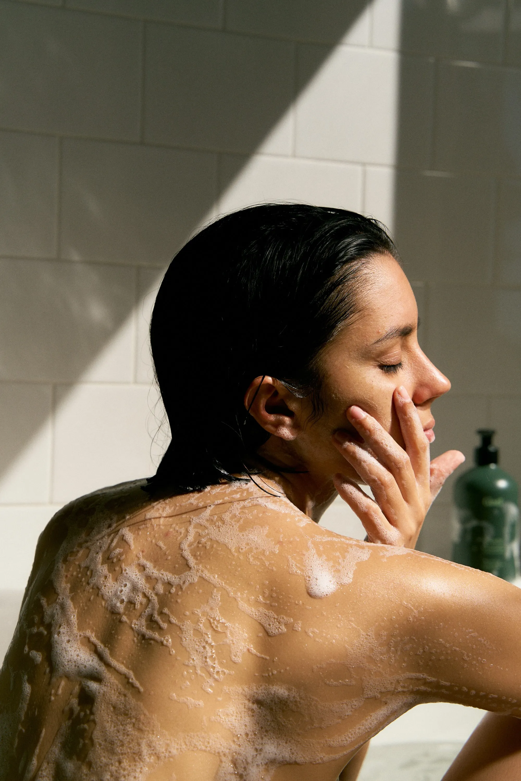 Woman with wet hair lathering in a tiled shower beside a Gntl bottle