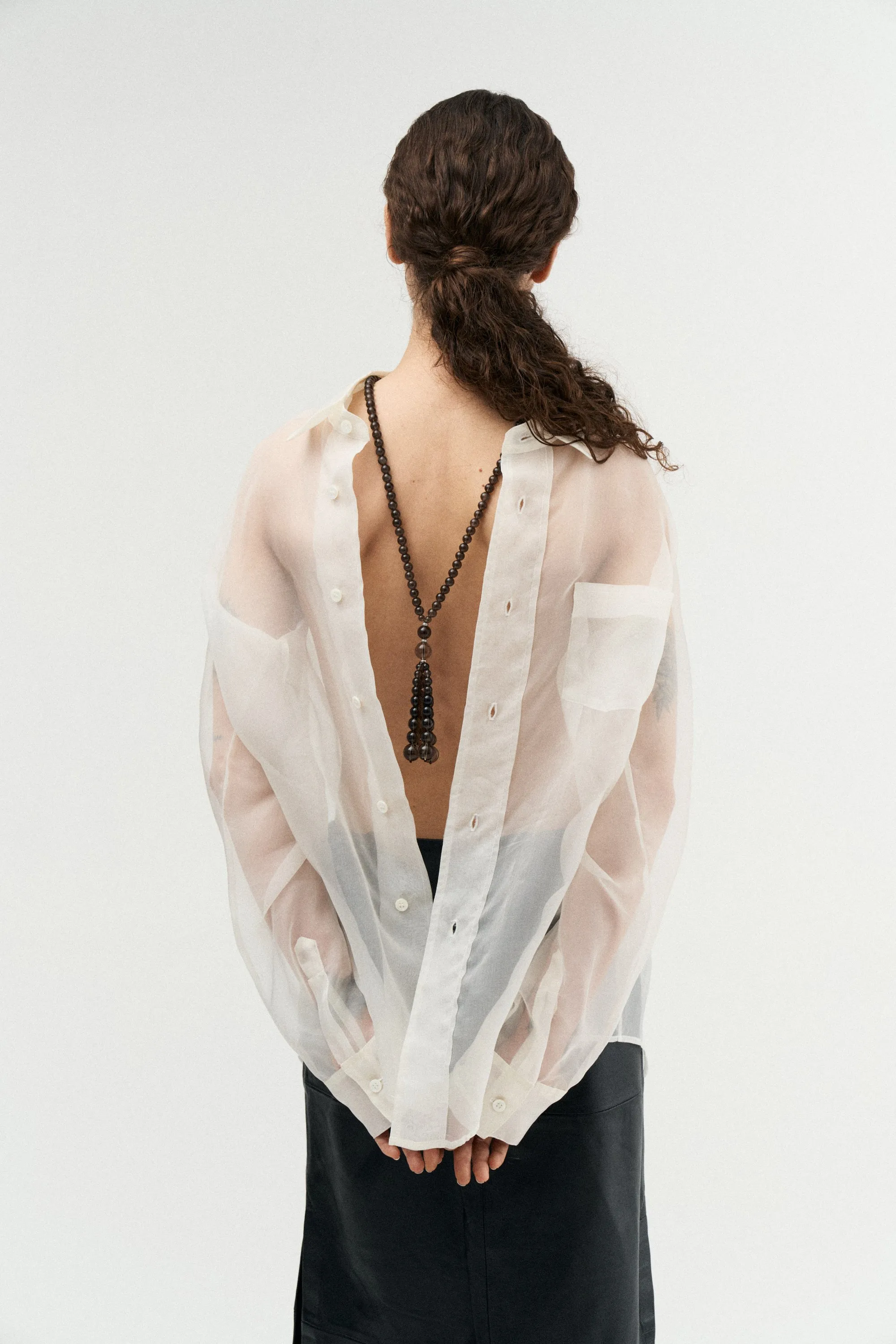 Model from behind in a sheer white shirt with a long beaded tassel necklace down her back