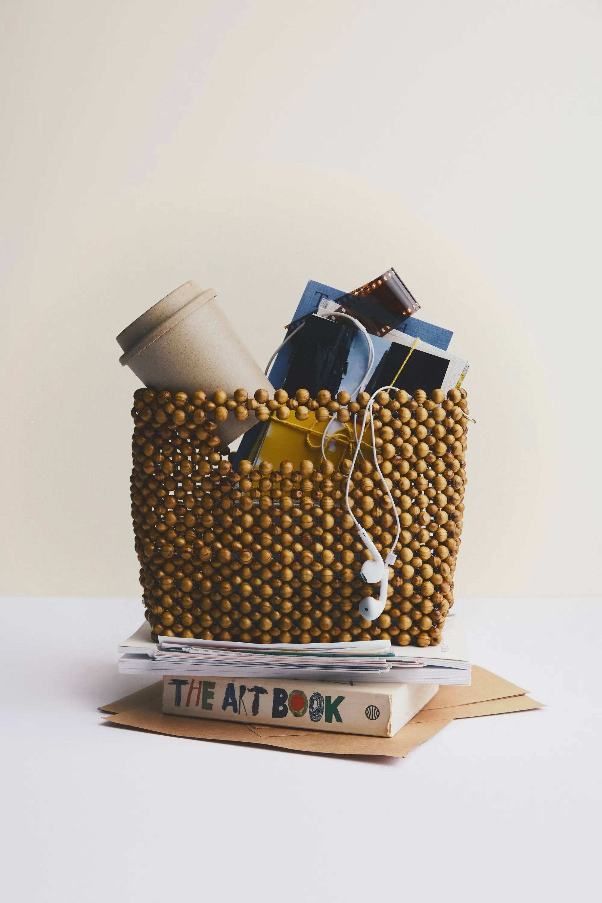 Beaded basket bag filled with a coffee cup, film photos and earphones, on stacked books and envelopes