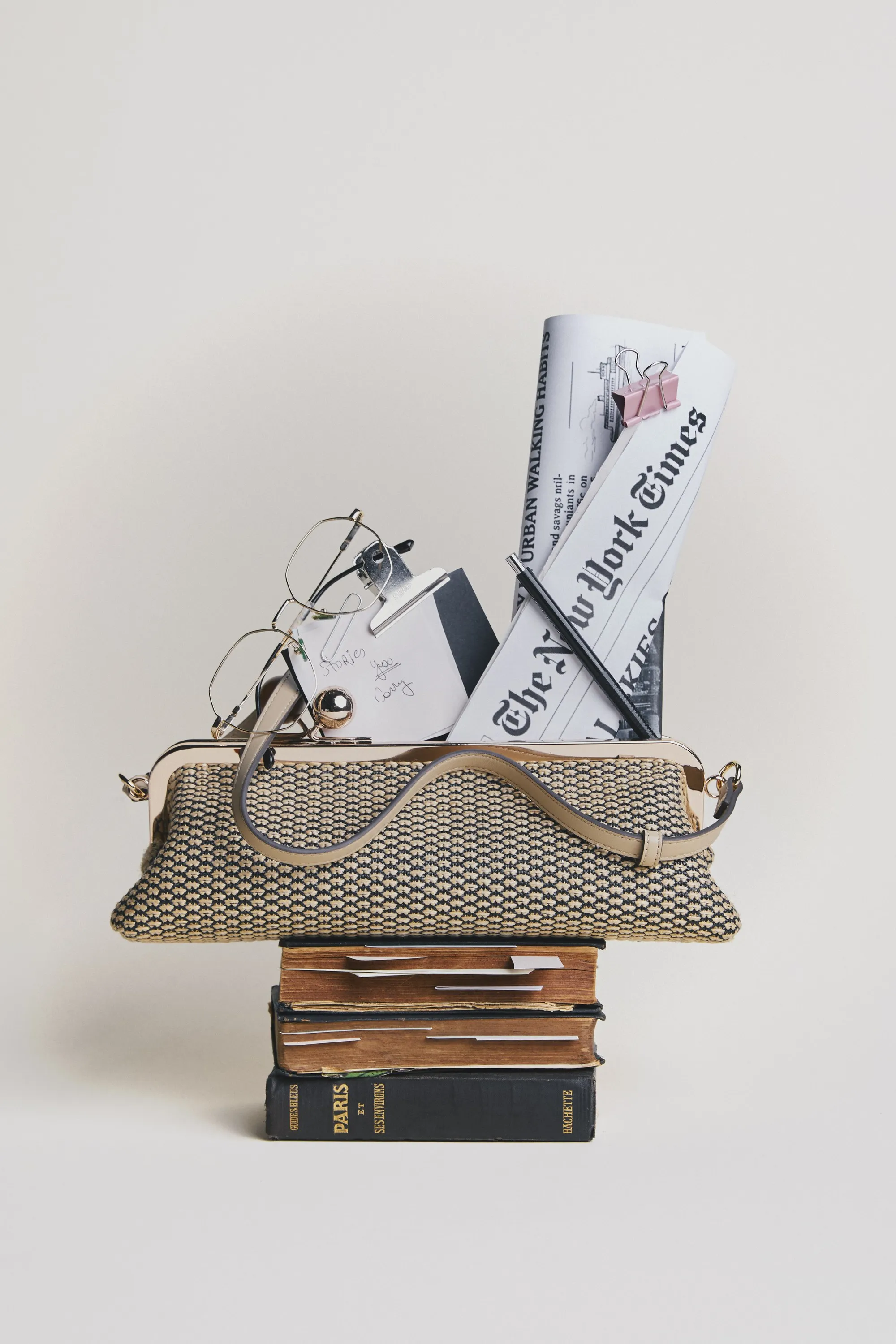 Woven frame bag with reading glasses and The New York Times, on a stack of old books