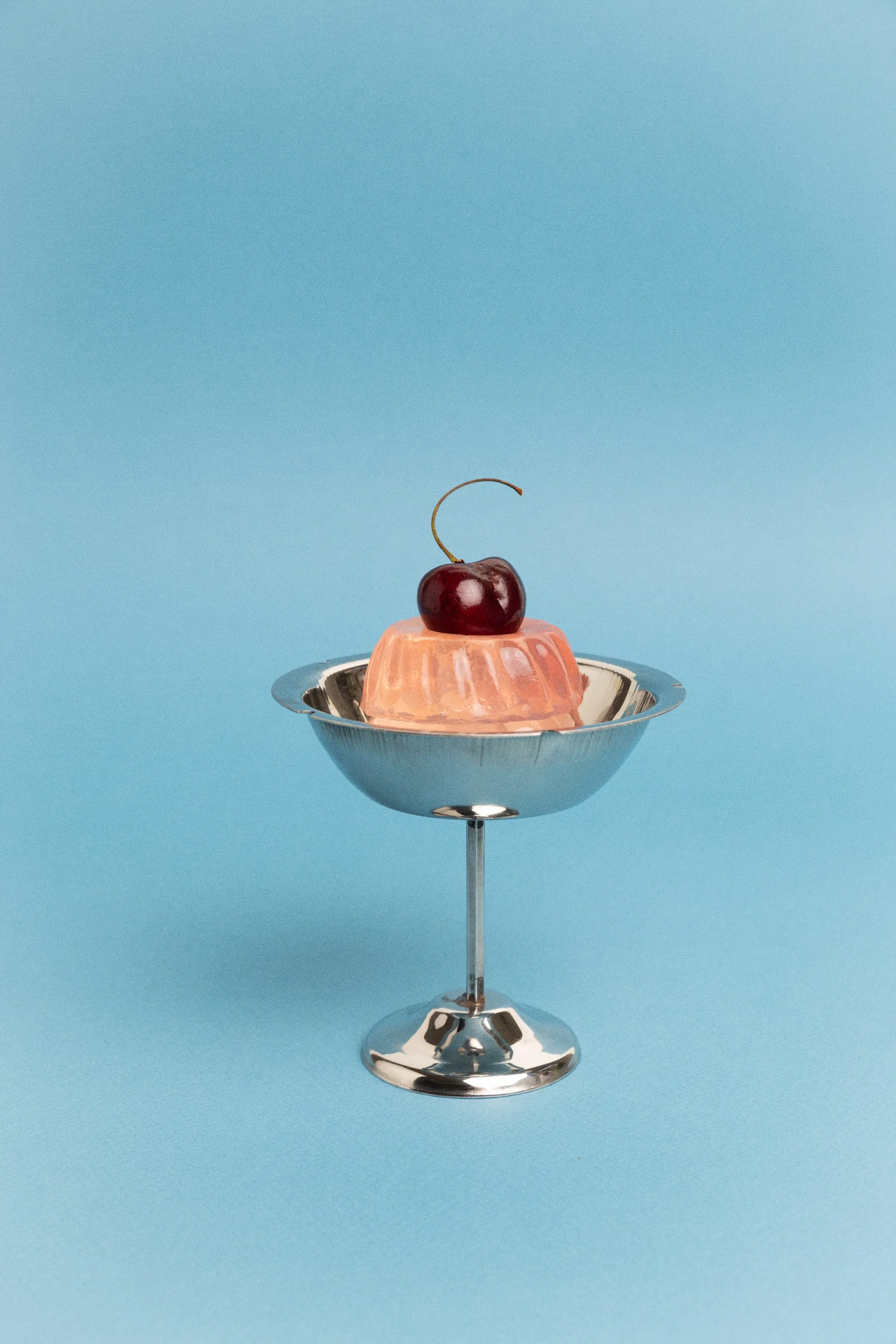 Peach jelly dessert topped with a cherry in a silver sundae cup, on pale blue