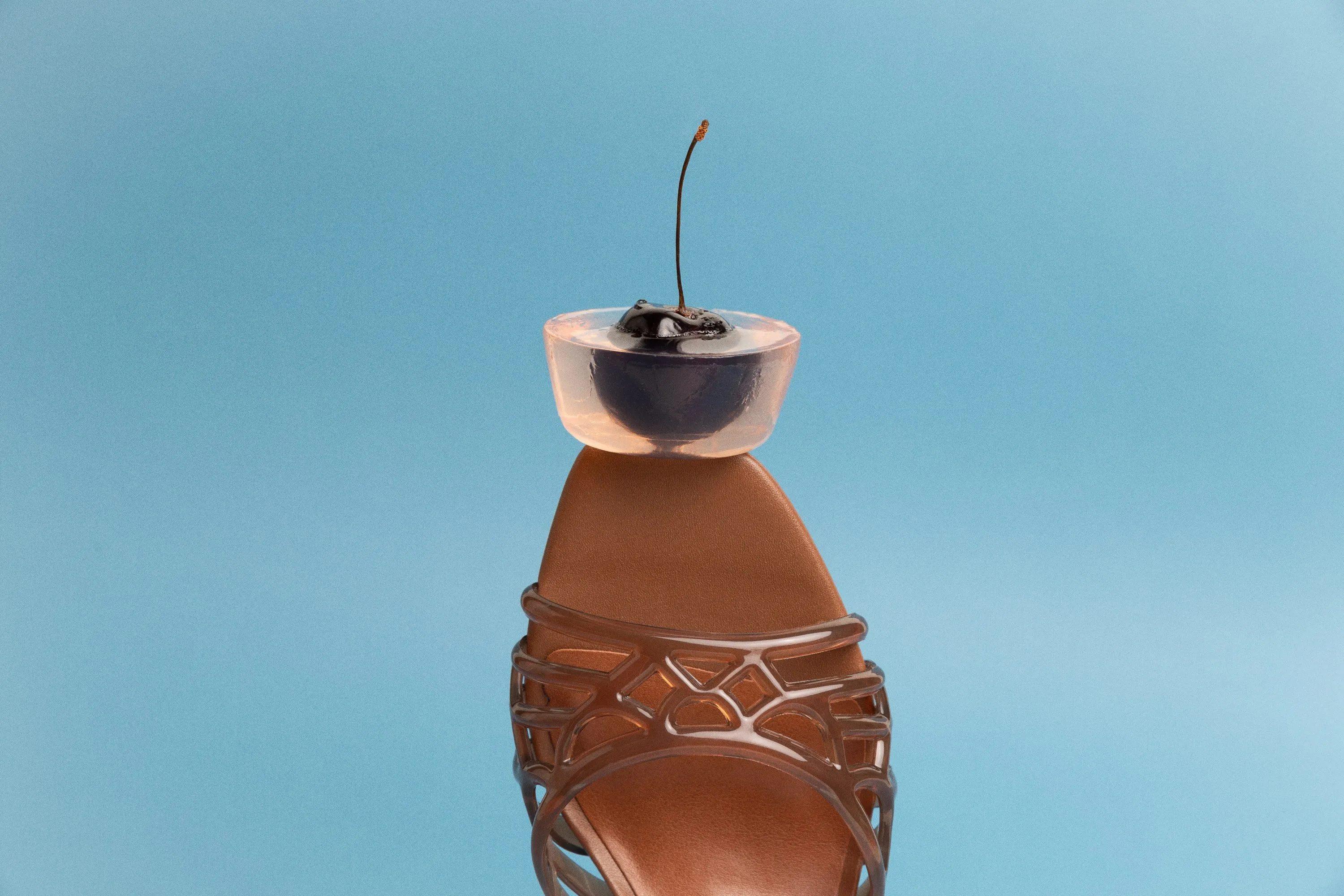 Cherry in a clear jelly cup balanced on the toe of a brown Dolce Vita sandal, on pale blue