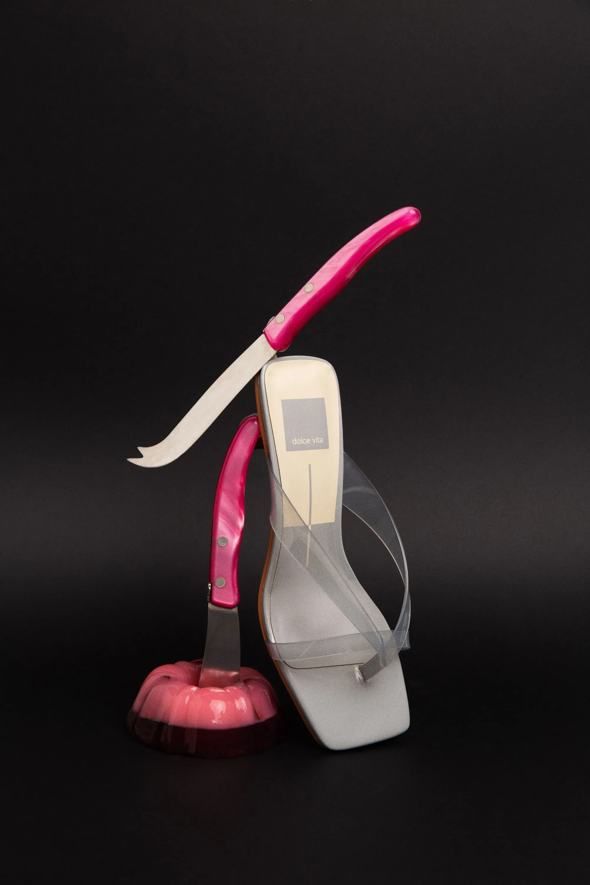 Clear Dolce Vita thong sandal balanced with pink cheese knives and a jelly mold, on black
