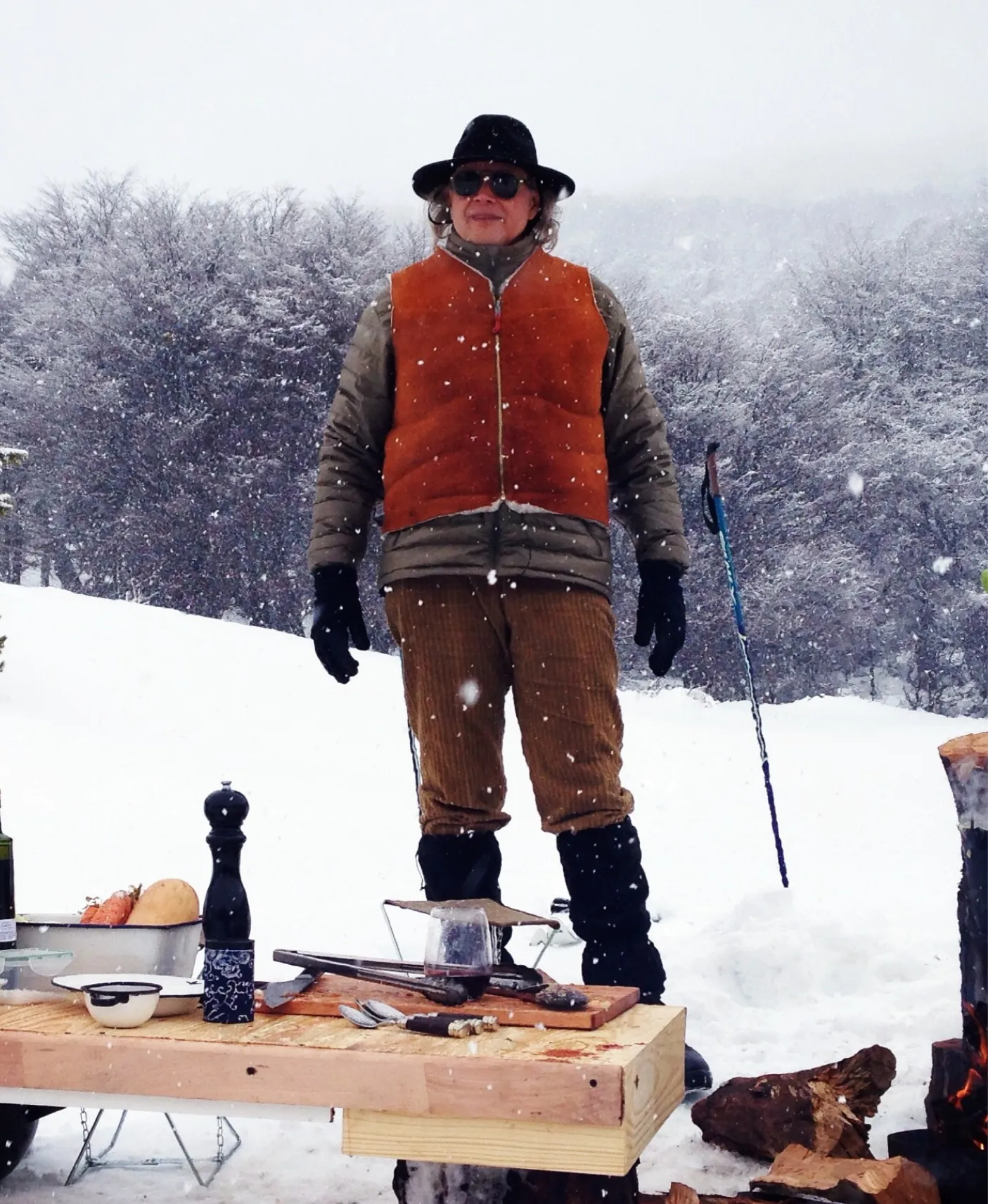 Francis Mallmann in a hat and suede vest standing in falling snow behind an outdoor cooking table in Patagonia