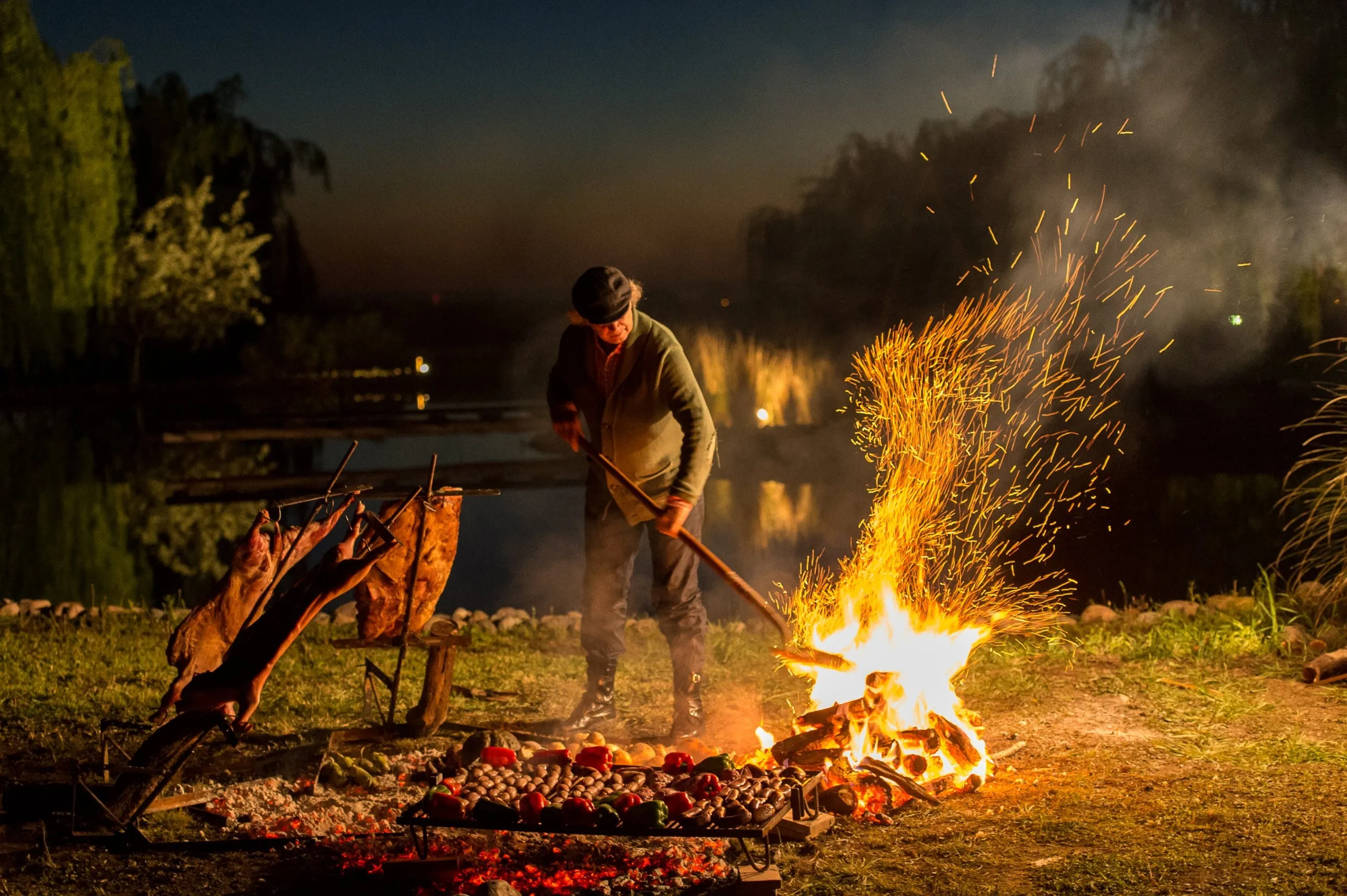 Francis Mallmann tending an open fire at night, sparks rising over a grill of vegetables and lamb on the asado cross