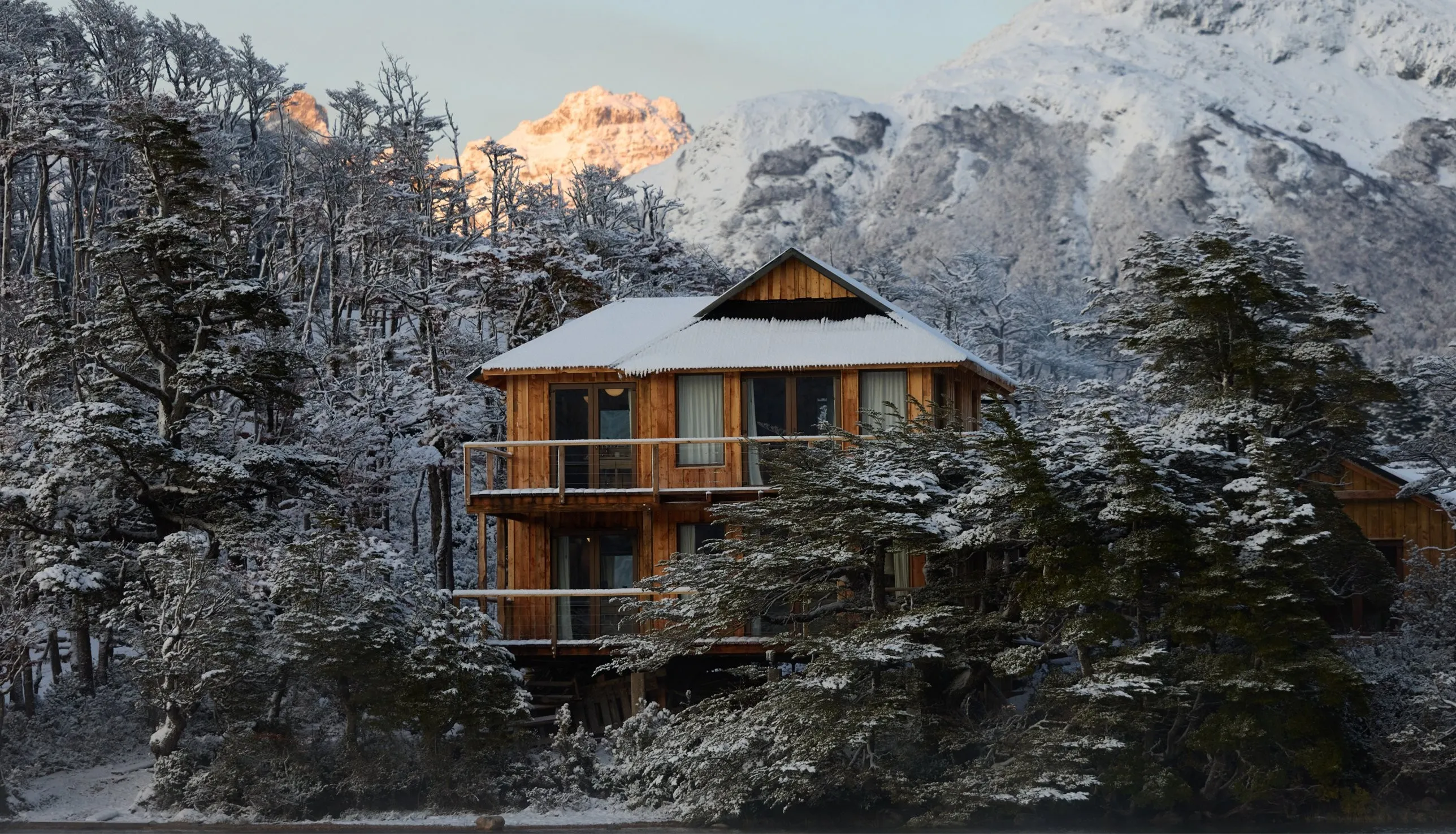Wooden La Isla lodge among snow-covered trees, a peak lit pink at dawn behind