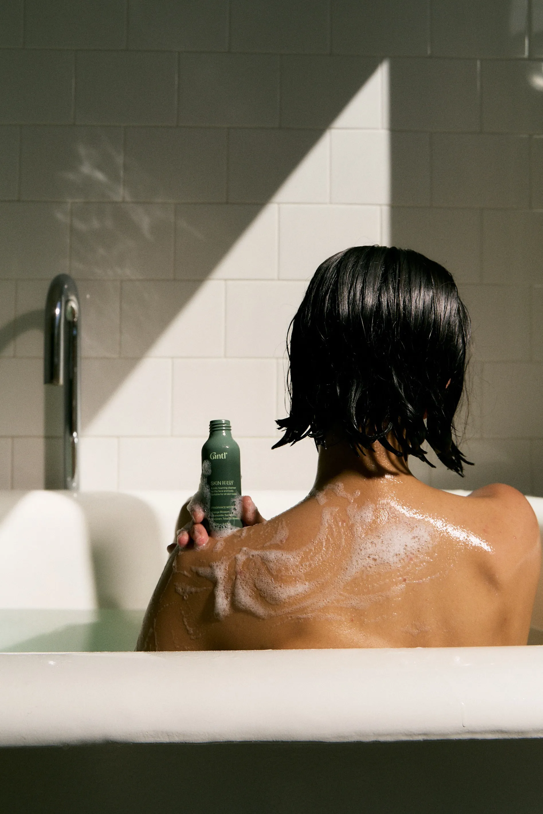 Woman seen from behind in a bath holding a small Gntl bottle